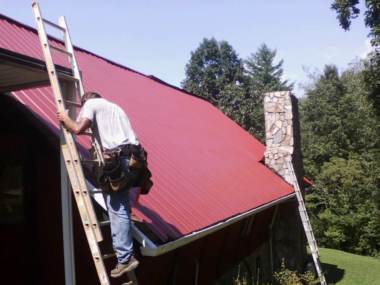 Roofer on a ladder beside a red metal roof and stone chimney — Soffit & Fascia Repair by Roofing Brothers in Pattersonville